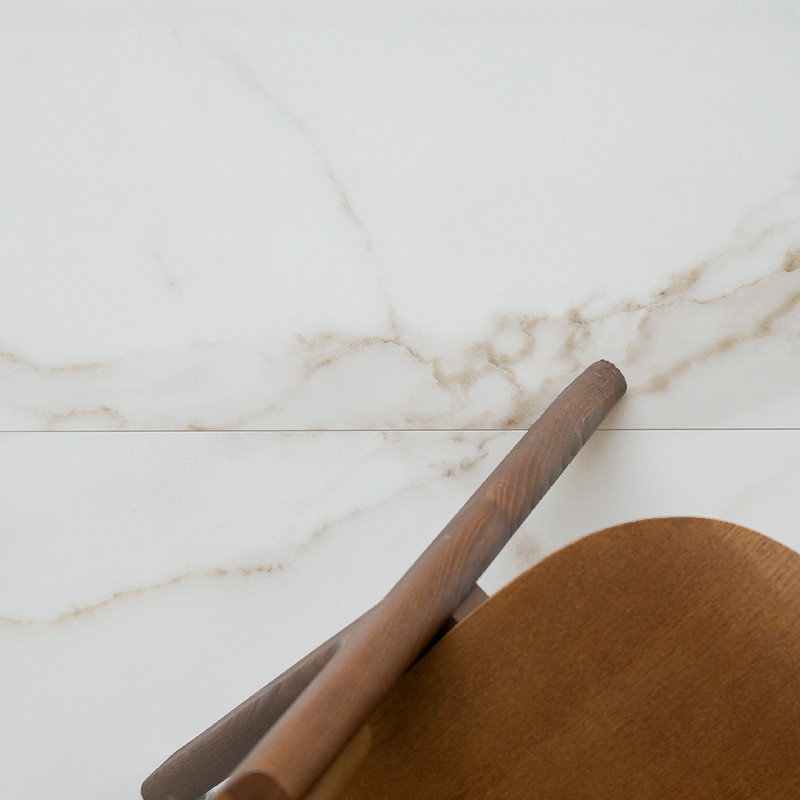 Silla de madera parcialmente visible, apoyada contra una pared de mármol blanco pulido con sutiles vetas beige, en un ambiente m