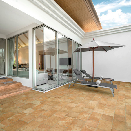 Deux chaises longues grises reposent sous un grand parasol sur un patio carrelé de couleur beige. Adjacente, une pièce moderne a
