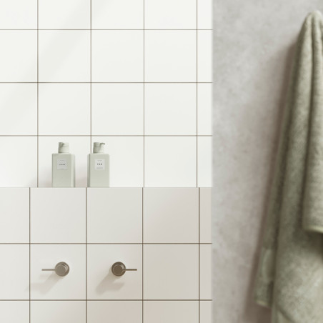 White tiles form a bathroom wall with two cylindrical handles below two bottles labeled `HAIR` and `BODY.` A green towel hangs n