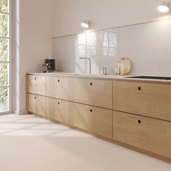 A modern kitchen features light wooden cabinets against white tiled walls. A coffee maker and cutting board sit on the counter. 