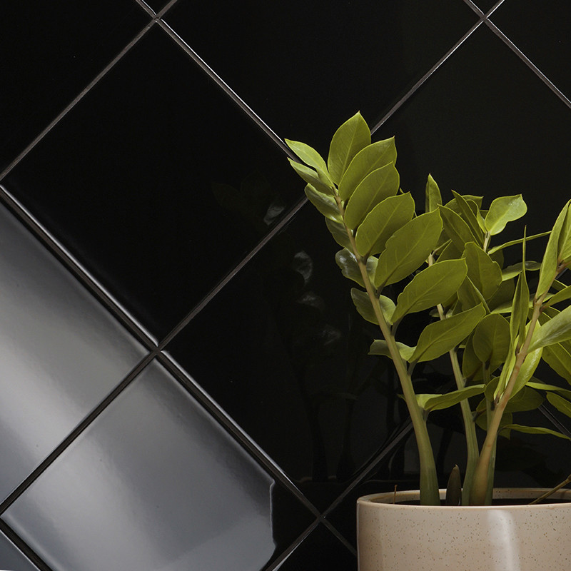 Ladrilhos pretos brilhantes formam um padrão diagonal na parede, destacando sua superfície elegante. Um vaso de planta com folha