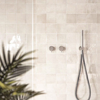 Glänzende beige Fliesen säumen eine Badezimmerwand und reflektieren das Licht. Ein Duschkopf und Bedienelemente sind montiert, m