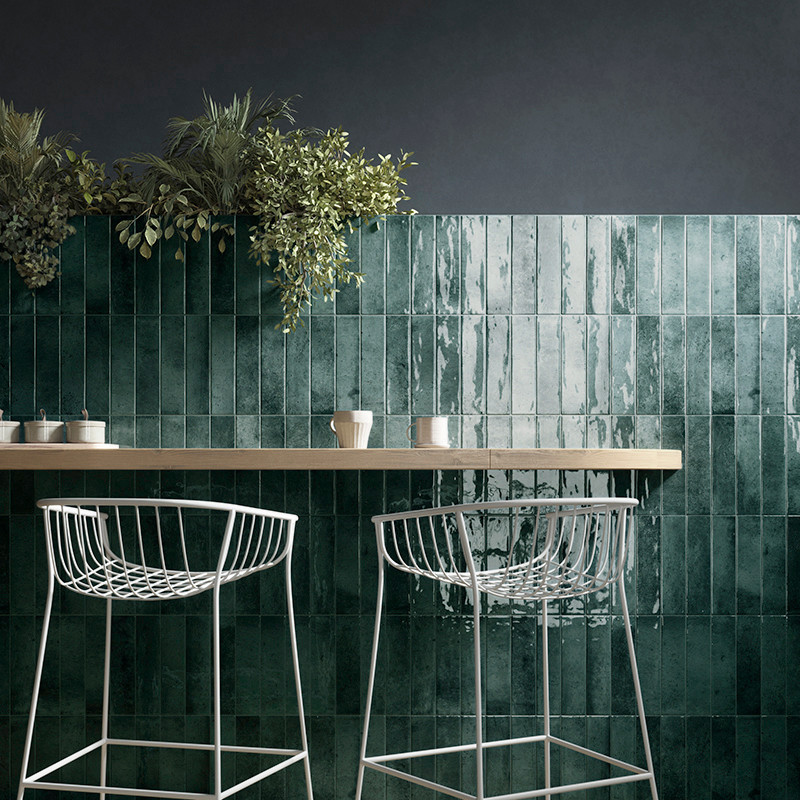 Des carreaux verts et brillants forment une toile de fond murale sous des plantes suspendues. Des tabourets de bar en métal blan
