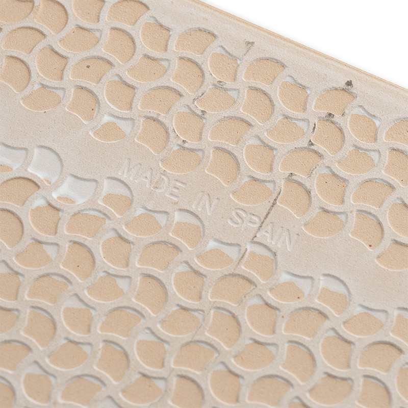 Un azulejo beige presenta un patrón repetido de formas pequeñas y curvas. El texto en relieve dice `MADE IN SPAIN`, agregando de