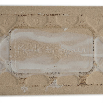 Um ladrilho retangular bege exibe o texto em relevo “Made in Spain” cercado por um padrão texturizado com os números 9, 5, 7 aci