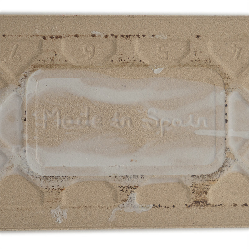 Eine rechteckige beige Fliese zeigt den geprägten Text „Made in Spain“, umgeben von einem strukturierten Muster mit den Zahlen 9