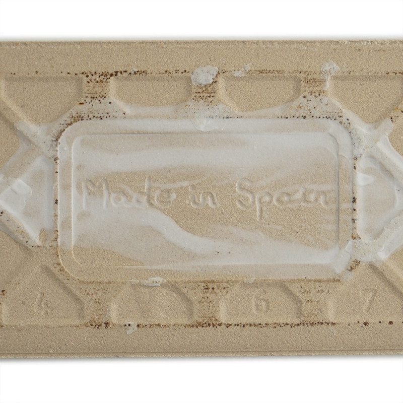 Een rechthoekige beige tegel heeft de opdruk `Made in Spain` in het midden, omgeven door verhoogde geometrische patronen en cijf