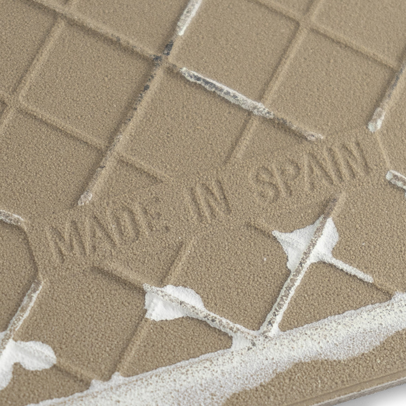 Un azulejo cerámico beige de textura rugosa muestra el texto `MADE IN SPAIN`. Presenta un patrón de cuadrícula con restos de lec
