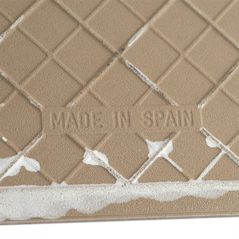 El azulejo presenta una superficie beige con diseño de rombos y ligeras marcas blancas. El texto dice `MADE IN SPAIN` grabado en
