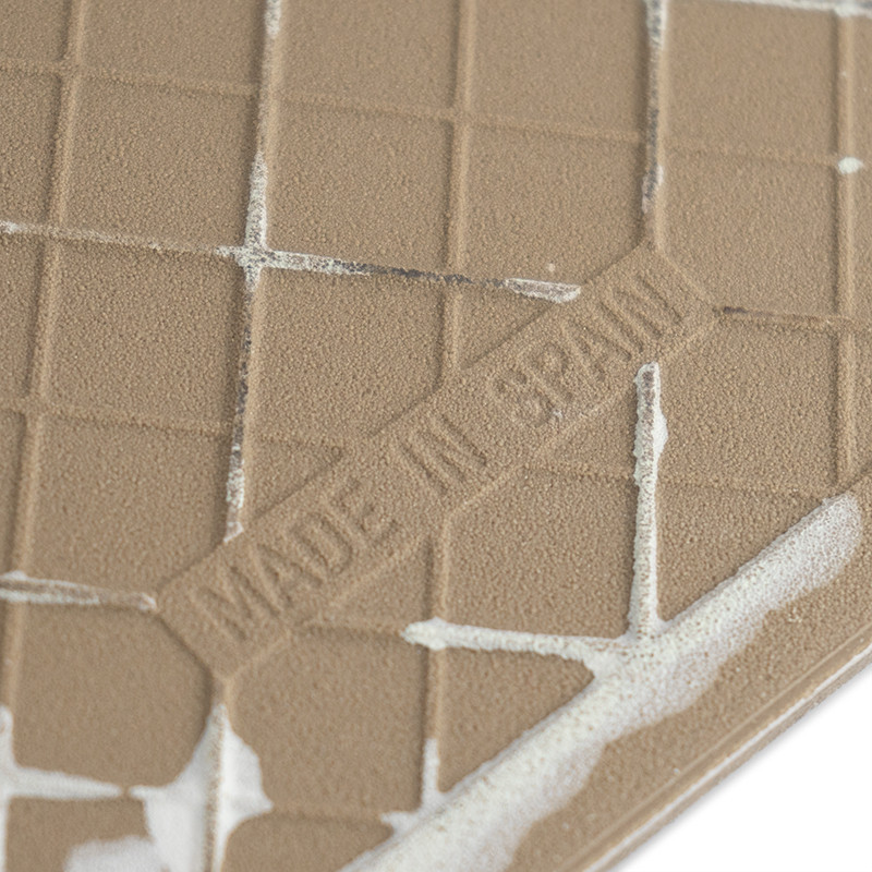 Un azulejo marrón texturizado, grabado con `MADE IN SPAIN`, muestra un patrón de cuadrícula. Está parcialmente cubierto con adhe