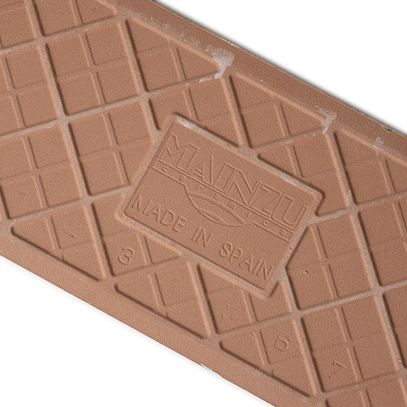 A rectangular, brown tile with a grid pattern embossed. It features a central logo: `MAINZU` and `MADE IN SPAIN,` set diagonally