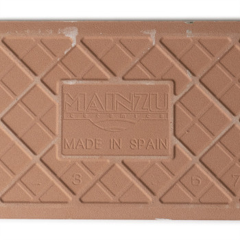 Eine strukturierte, braune Keramikfliese zeigt ein Gittermuster mit dem geprägten Text „MAINZU CERAMIC MADE IN SPAIN“ und den Za