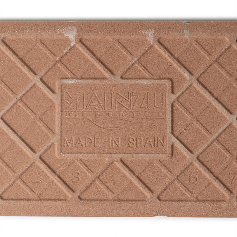 Eine strukturierte, braune Keramikfliese zeigt ein Gittermuster mit dem geprägten Text „MAINZU CERAMIC MADE IN SPAIN“ und den Za