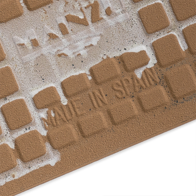 The tile displays a grid of small, raised squares. Faded text reads `MADE IN SPAIN,` surrounded by rough, textured beige materia