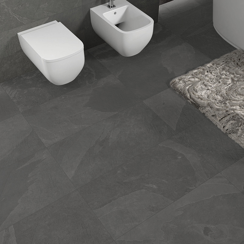 Gray tiles cover the bathroom floor and walls, supporting a wall-mounted toilet and bidet. A patterned rug lies nearby.