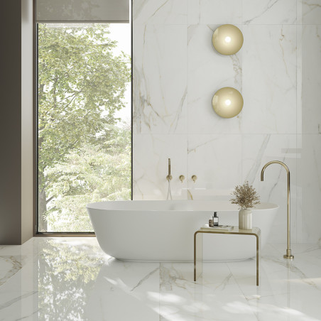 Een vrijstaand wit bad rust op een glanzende marmeren tegelvloer, omgeven door natuurlijk licht door een groot raam en een minim