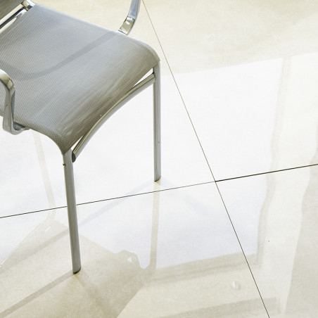 A metallic chair with a gray seat casts a shadow on a glossy, light-colored tiled floor illuminated by natural light.