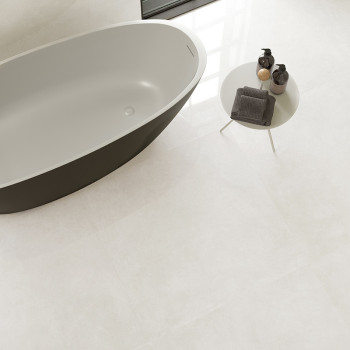 A modern, oval bathtub sits beside a round, white table with toiletries in a minimalistic bathroom with light-colored tiles.