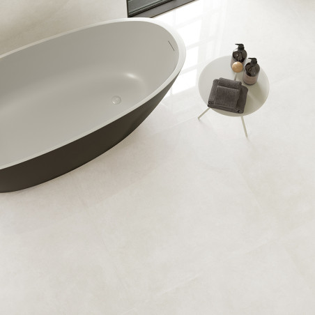 A modern, oval bathtub sits beside a round, white table with toiletries in a minimalistic bathroom with light-colored tiles.