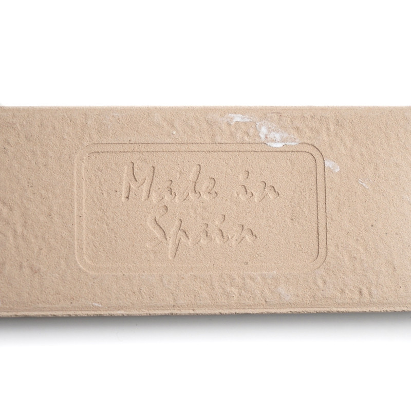 Um ladrilho retangular bege exibe o texto em relevo “Made in Spain” no centro, com uma textura áspera e sutis manchas brancas.
