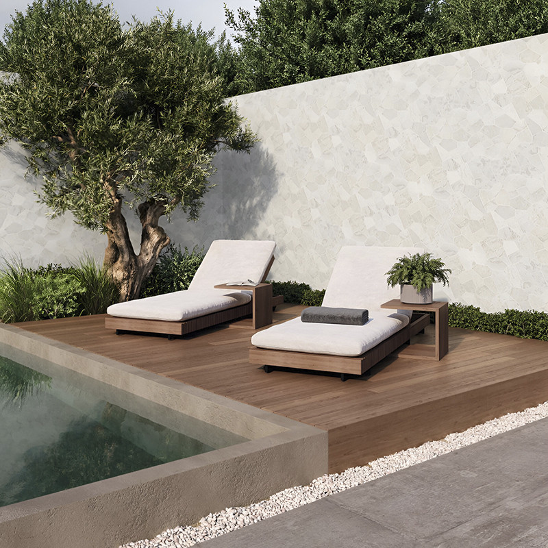 Zwei gepolsterte Liegestühle stehen auf einer Holzterrasse neben einem Pool, umgeben von üppigem Grün und einer Steinmauer.
