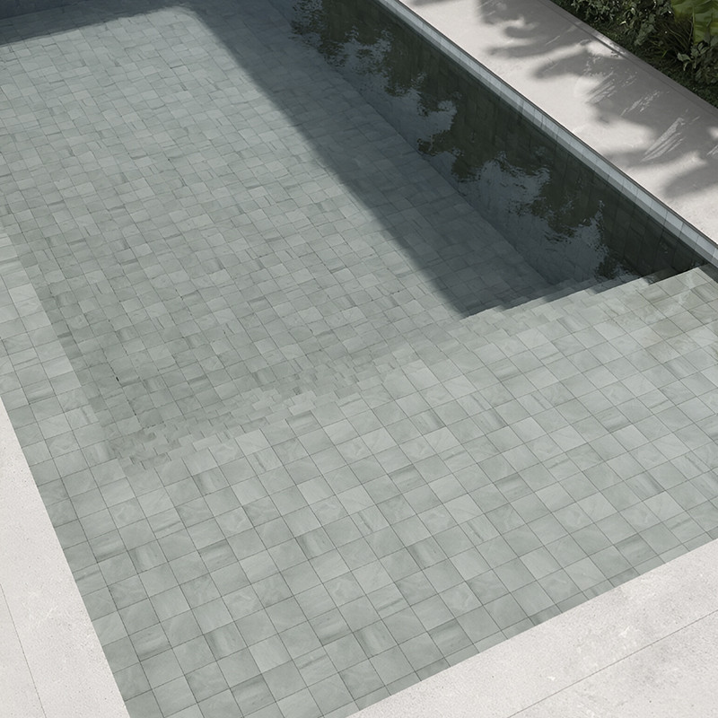 Light gray stone tiles form a submerged pool floor, reflecting sunlight, surrounded by concrete edges and green foliage.