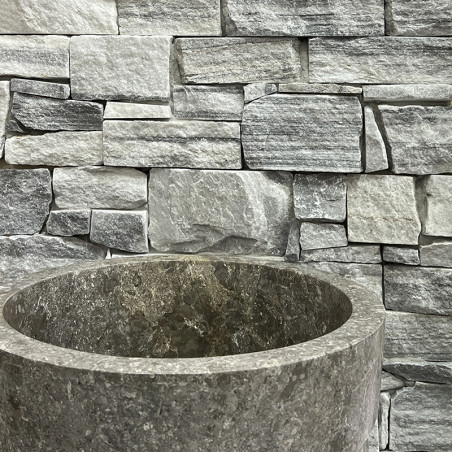 Round stone basin sits in front of stacked, uneven rectangular gray stone tiles, creating a rustic, textured backdrop.