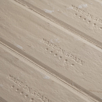 Un carreau beige affiche le texte « MADE IN SPAIN » estampé à plusieurs reprises, sur un fond texturé avec de subtiles lignes ho