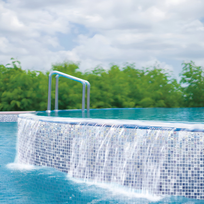 Wasser kaskadiert über den Rand eines Infinity-Pools mit Glasmosaik, vor üppigem Grün und einem bewölkten Himmel.