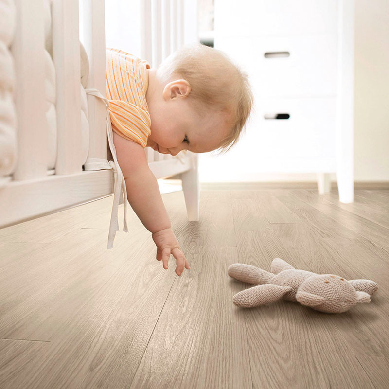 Ein Baby beugt sich über ein weißes Kinderbett und greift in einem sanft beleuchteten Raum nach einem Plüschhasen auf einem Holz