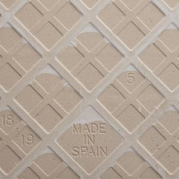 De tegel heeft een beige, diagonaal rasterpatroon met cijfers in reliëf. `MADE IN SPAIN` staat prominent in het midden gestempel