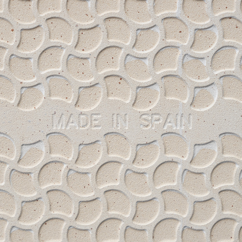 Tile features embossed, overlapping semicircular pattern. `MADE IN SPAIN` inscribed in center. Background is a light beige color