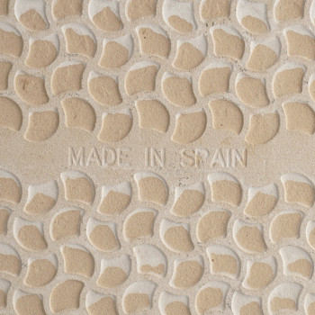 De beige tegel heeft een verhoogde, kiezelachtige textuur met `MADE IN SPAIN` in reliëf in het midden, wat een decoratief tintje