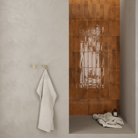 Eine glänzende, rotbraun geflieste Wand bildet einen Duschbereich mit einem Handtuch, das über einen Haken an einer glatten, hel