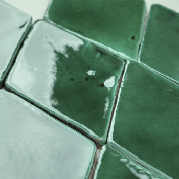 Grün glasierte Terrakottafliesen, die Licht reflektieren und in einem Fischgrätenmuster auf einer ebenen Fläche angeordnet sind.