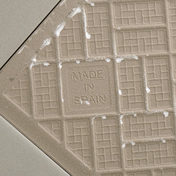 A beige ceramic tile features a grid pattern with the text `MADE IN SPAIN` at the center, surrounded by light, diagonal grout li