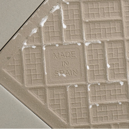 A beige ceramic tile features a grid pattern with the text `MADE IN SPAIN` at the center, surrounded by light, diagonal grout li