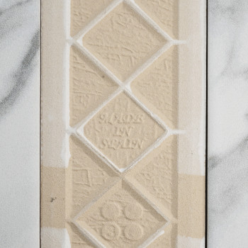 Un carreau beige texturé présente des losanges en relief avec le texte « MADE IN SWEDEN » au centre. Il repose sur un fond à mot