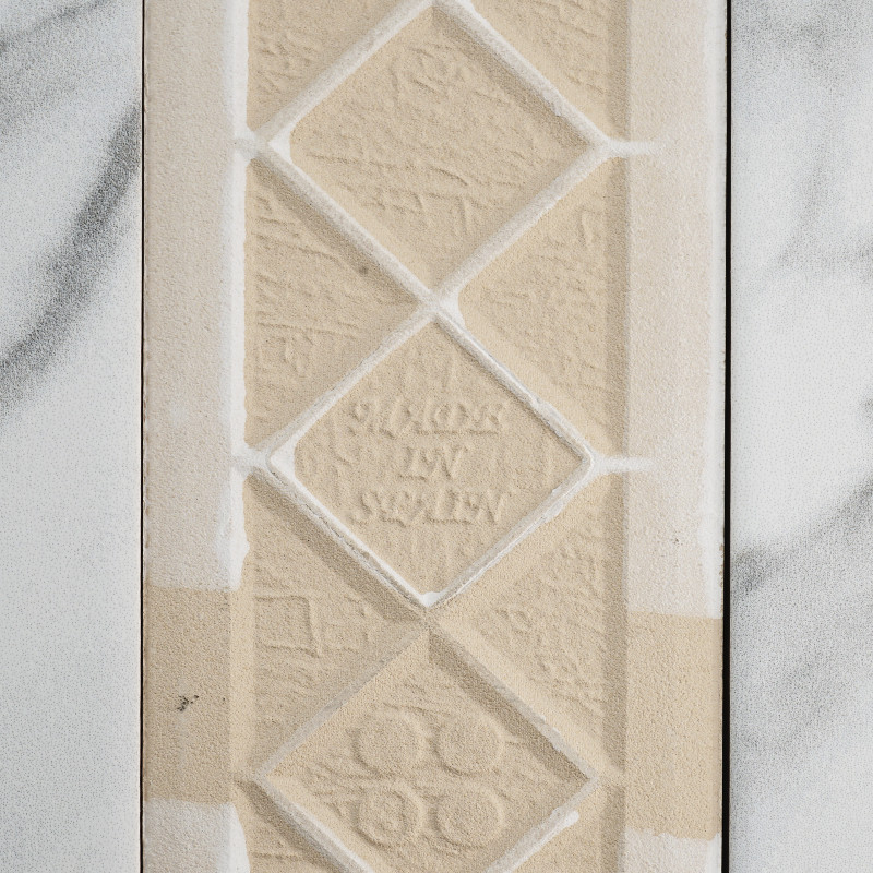 Un azulejo texturizado de color beige muestra formas de diamantes en relieve con el texto `MADE IN SWEDEN` en el centro. Se apoy
