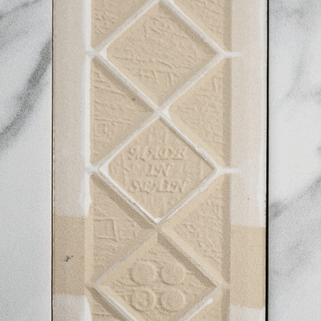 Un carreau beige texturé présente des losanges en relief avec le texte « MADE IN SWEDEN » au centre. Il repose sur un fond à mot