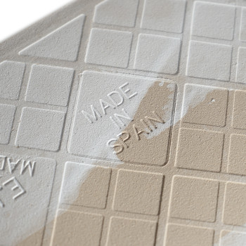 Auf einer hellbeigen Kachel ist inmitten eines Gittermusters der geprägte Text „MADE IN SPAIN“ zu sehen, der teilweise von einer