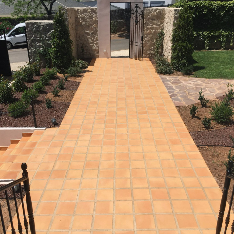 Terracotta tiles forming a wide walkway to an iron gate; flanked by flowerbeds with shrubs, a stone wall, steps with a railing and grass.