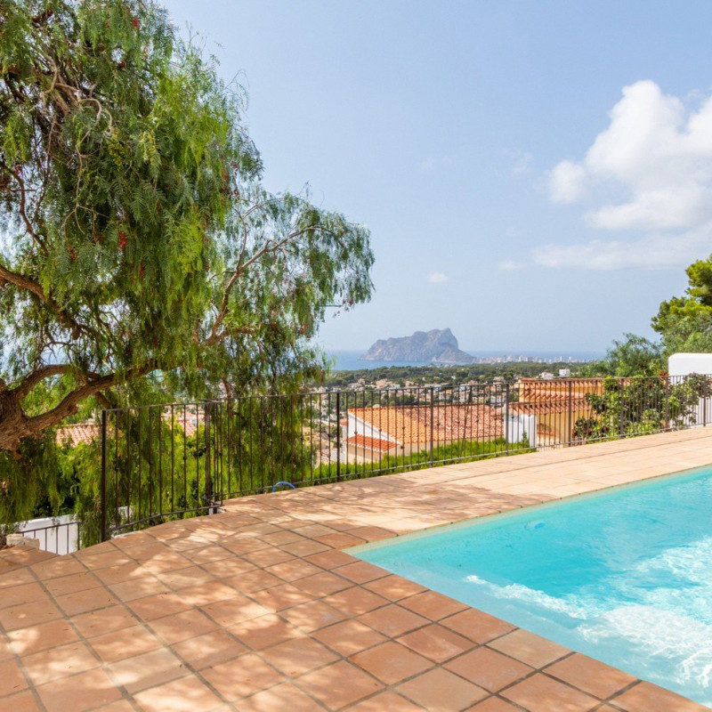 Losas de terracota junto a piscina rectangular; barandilla metálica y árbol colgante al borde; vistas al mar y un peñón rocoso sobre un barrio de casas con tejados.