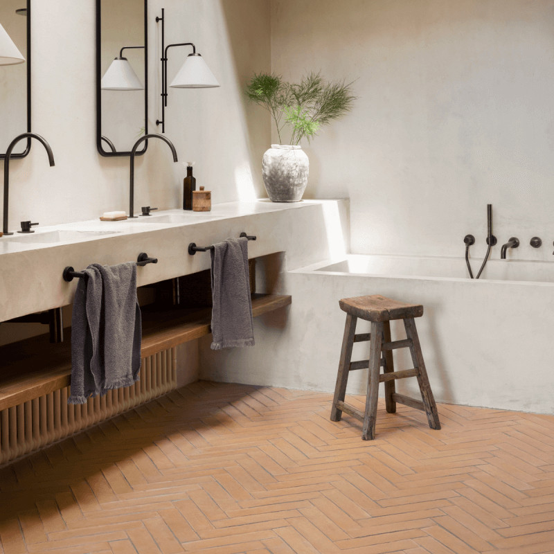 Los azulejos de terracota forman un patrón en espiga en el piso del baño, con un taburete de madera y una decoración minimalista