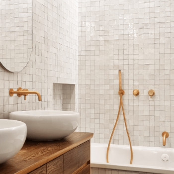 Uma pia oval branca fica sob uma torneira dourada, contra uma parede de azulejos quadrados brancos, criando uma estética moderna