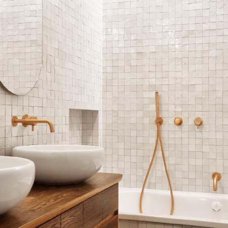 Uma pia oval branca fica sob uma torneira dourada, contra uma parede de azulejos quadrados brancos, criando uma estética moderna