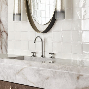 White terracotta tiles line a bathroom wall, reflecting light around a marble countertop with a modern faucet fixture.