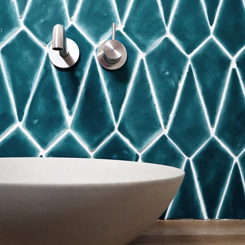 Blauwe geometrische terracottategels bedekken een badkamermuur boven een moderne witte wastafel, waarop twee metalen kranen zijn