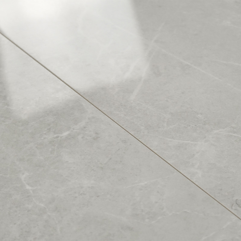 A smooth, light gray marble tile displays subtle veining and a reflective surface, laid flat with visible grout lines in a lit, 