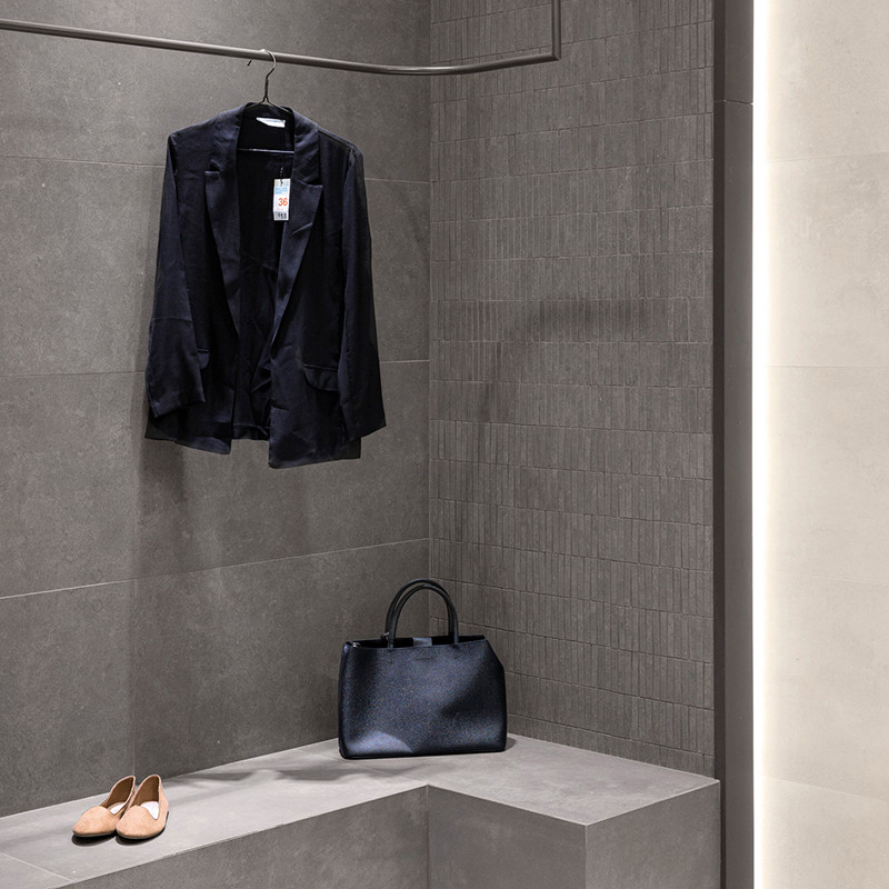 A black blazer hangs from a hook in a minimalist changing room with dark gray tiles, alongside a black handbag and beige shoes r